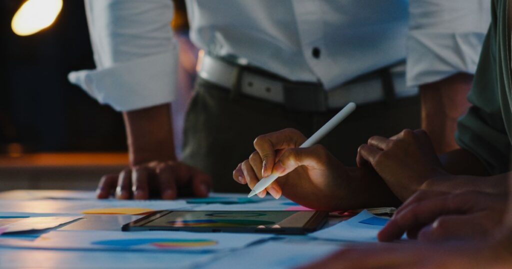 Two people collaborate, using a digital tablet and stylus amidst colorful charts and designs on a table.