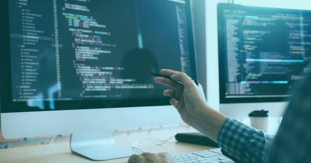 Person coding on dual monitors, focusing on programming software, with a coffee cup nearby.