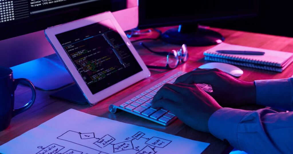 A person types on a keyboard connected to a tablet, surrounded by coding on multiple screens and a flowchart on paper.