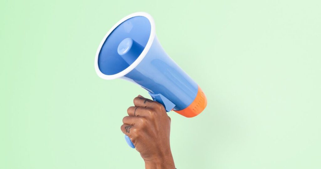A hand holding a blue megaphone against a pastel green background, symbolizing communication and public speaking.