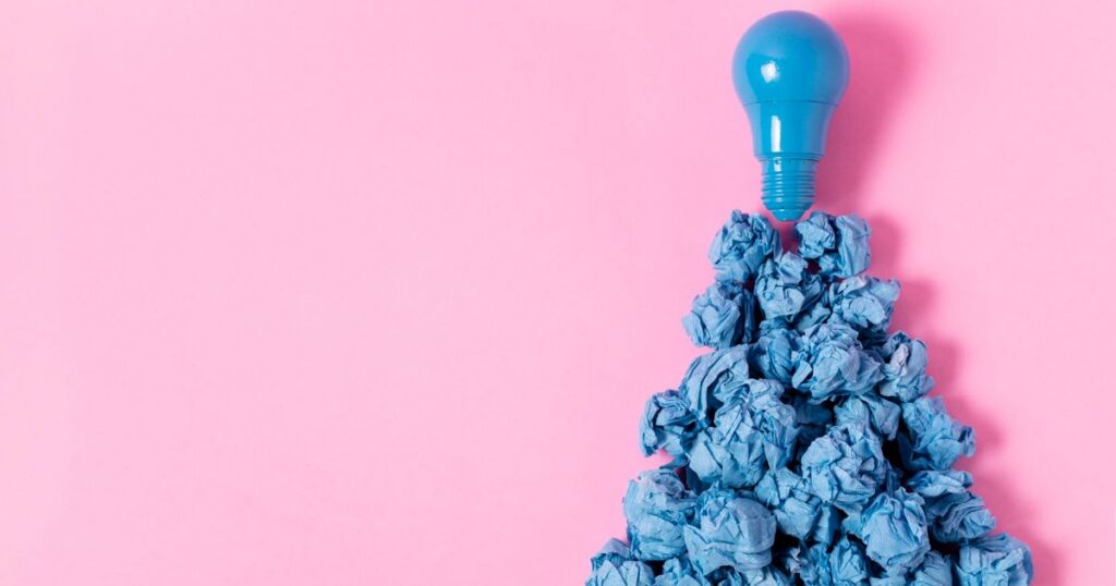 Blue crumpled paper forming a mound beneath a blue light bulb on a pink background, symbolizing creativity and ideas.
