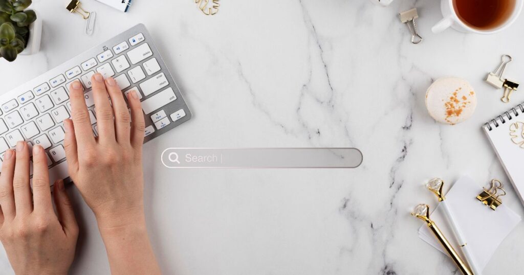 A hand typing on a keyboard next to a search bar on a marble desk with a cup, notepad, and office supplies.