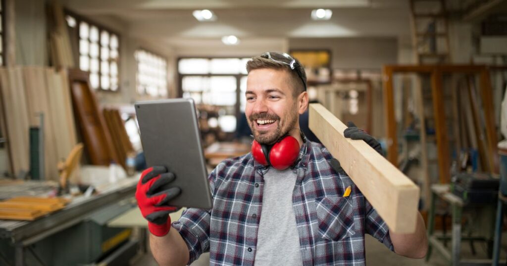 Carpenter in work gloves, wearing headphones, smiles while holding a wooden plank and a tablet in a workshop.