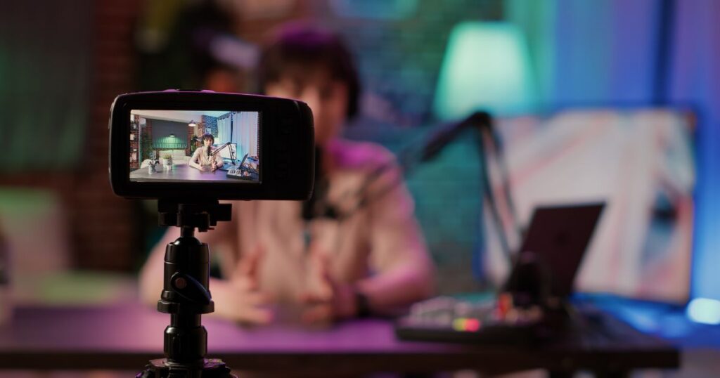 Person hosting a podcast, viewed through a camera lens, with colorful studio lighting and audio equipment in the background.